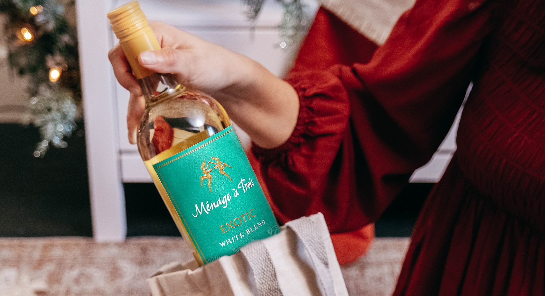 woman in a red dress pulling out a bottle of Menage a Trois exotic white blend from a gift bag. Get great bottles of wine to gift this season from One Stop Wine Shop.