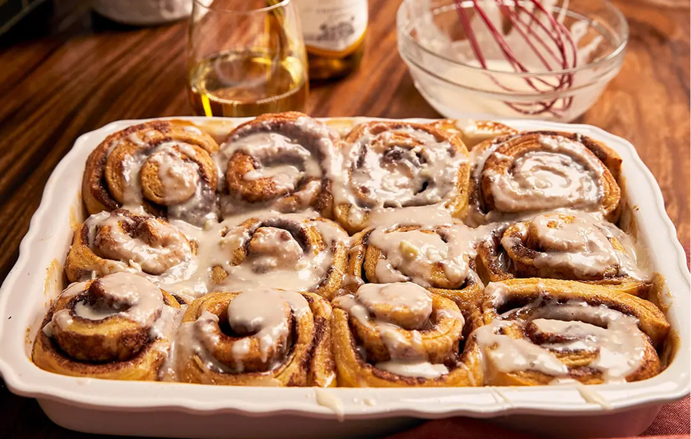 A tray of cinnamon rolls frosted with eggnog frosting with a bottle and glass of Sutter Home chardonnay in the background. Get this recipe and more from One Stop Wine Shop.