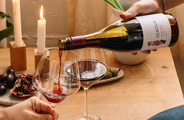 Hand pouring a glass of SEAGLASS pinot noir in front a table with olives, pomegranates, and lit white candles in modern wood holders. Shop this season favorites and more at One Stop Wine Shop