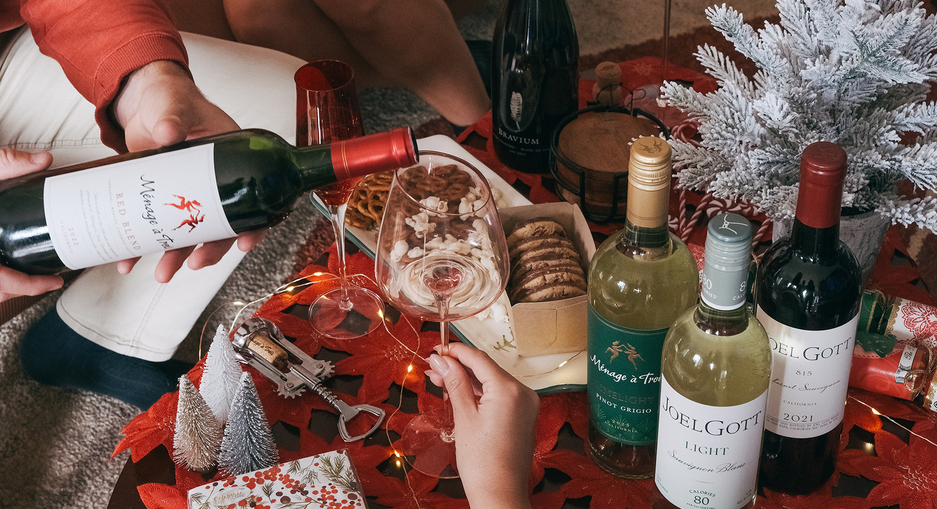 various bottles of wine on a holiday toable festooned with tiny trees, boxes of cookies and snacks, and a festive red tablecloth. Explore the One Stop Wine Shop wine gift guide.