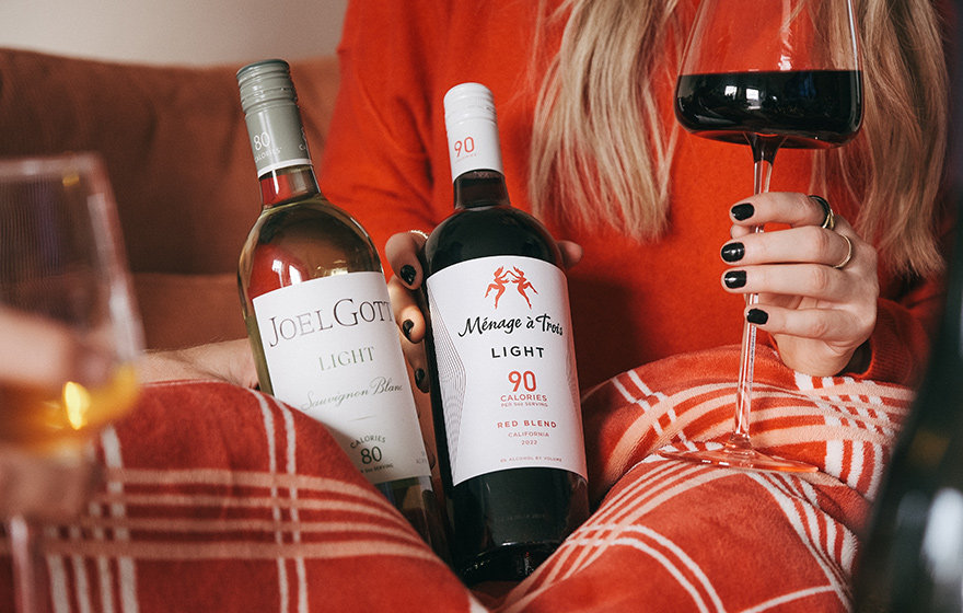 A women wearing a red sweater in a cozy blanket holding a glass of red wine and bottles of Joel Gott light sauvignon blanc and Menage a Trois light red blend wines.
