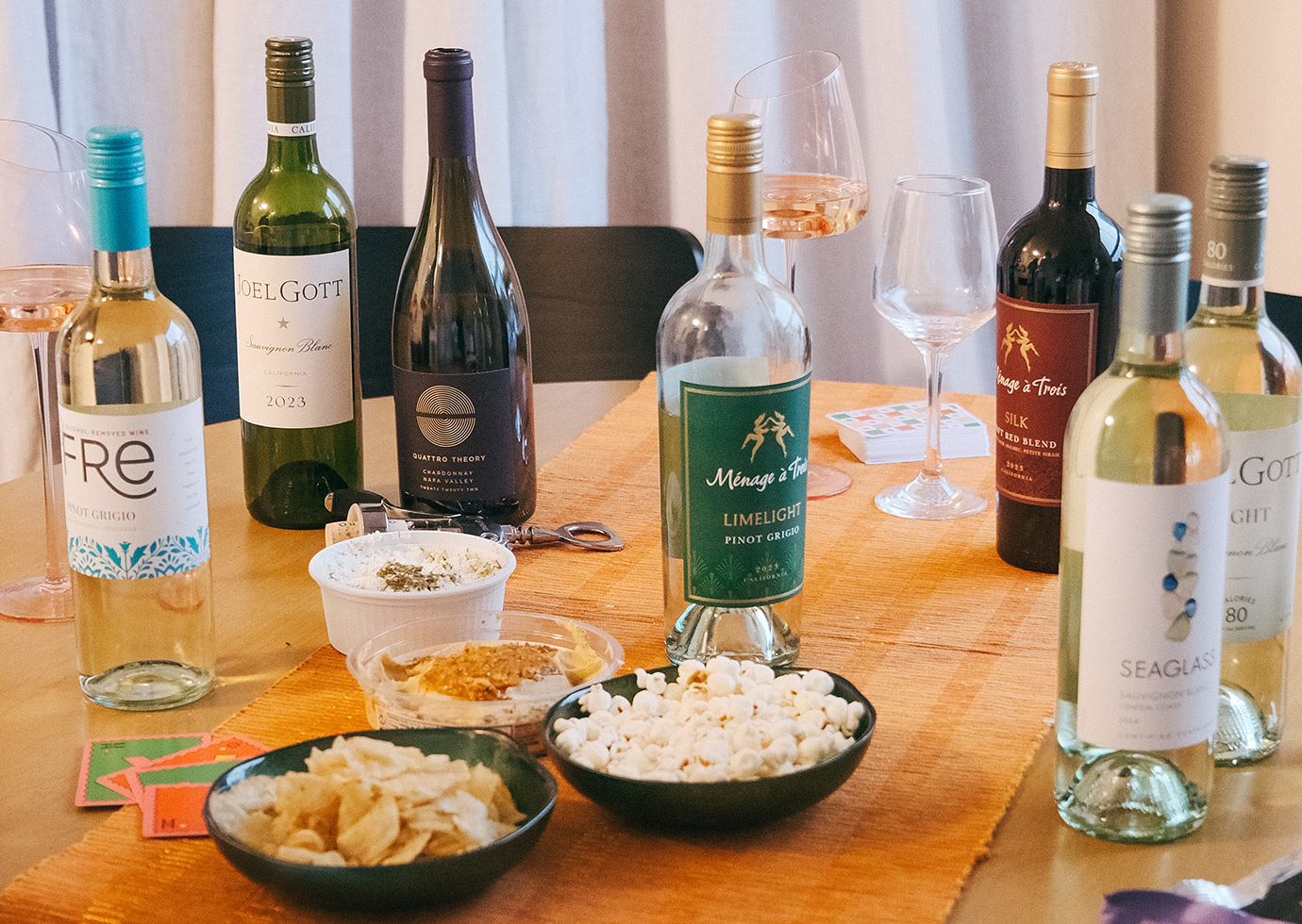 Various bottles of wines from One Stop Wine Shop on a dining table with bowls of snacks and cards. Take our wine recommender quiz to find your new favorite.