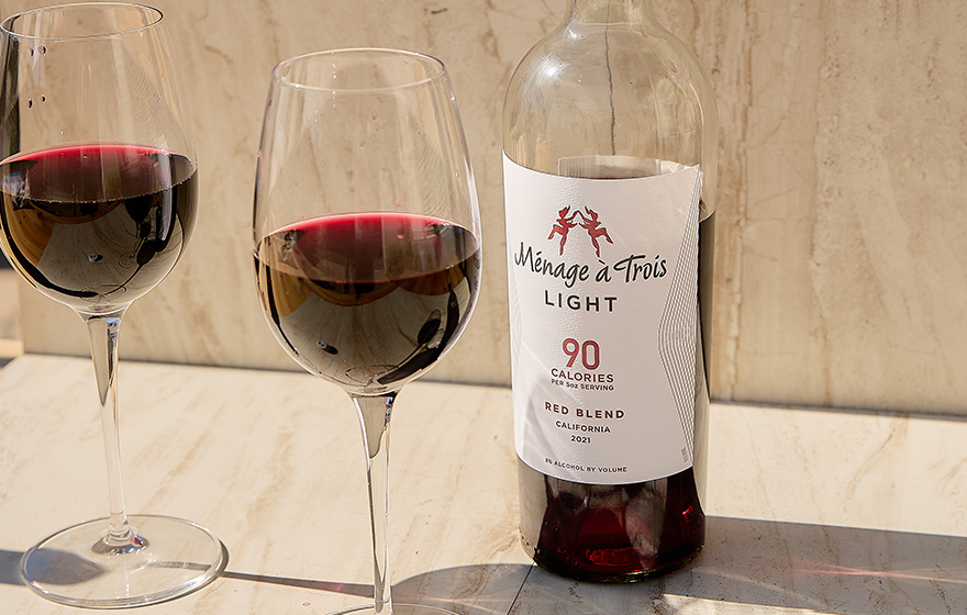 A bottle and two glasses of Menage a Trois light red blend, just 90 calories per 5 ounce serving. Buy this lower calorie wine and more at One Stop Wine Shop.