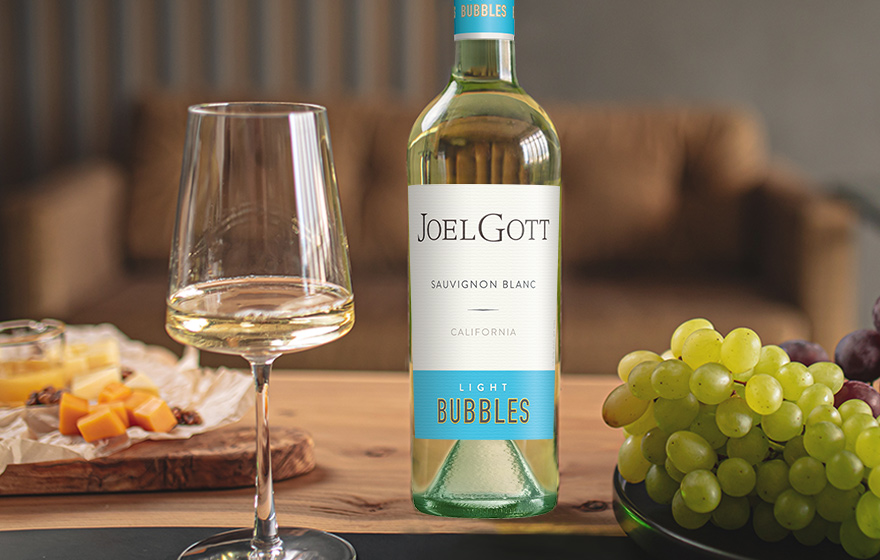 A bottle and glass of Joel Gott sauvignon blanc light bubbles on a table with grapes and cheese. Buy this new sparkling wine and more at One Stop Wine Shop.