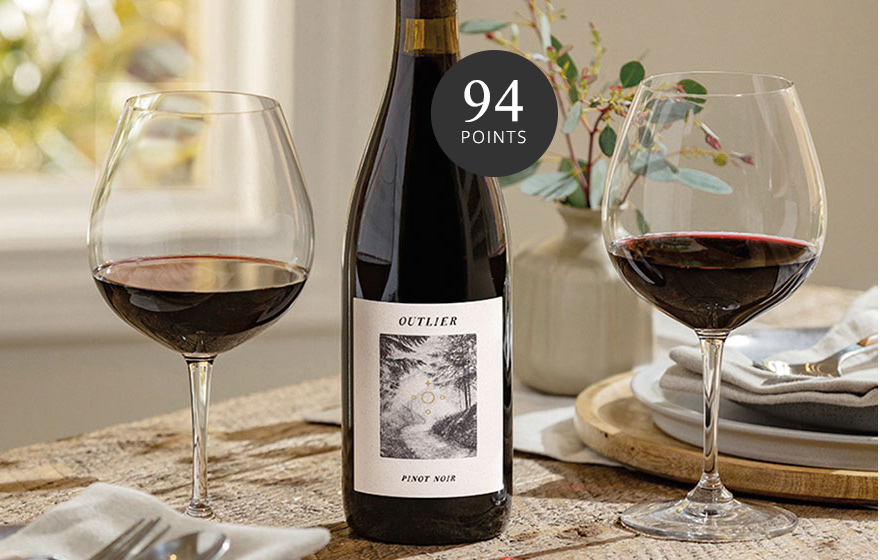 A bottle and two glasses of Outlier pinot noir on a wooden dinner table, rated 94 points. Buy this top-scoring wine and more at One Stop Wine Shop.