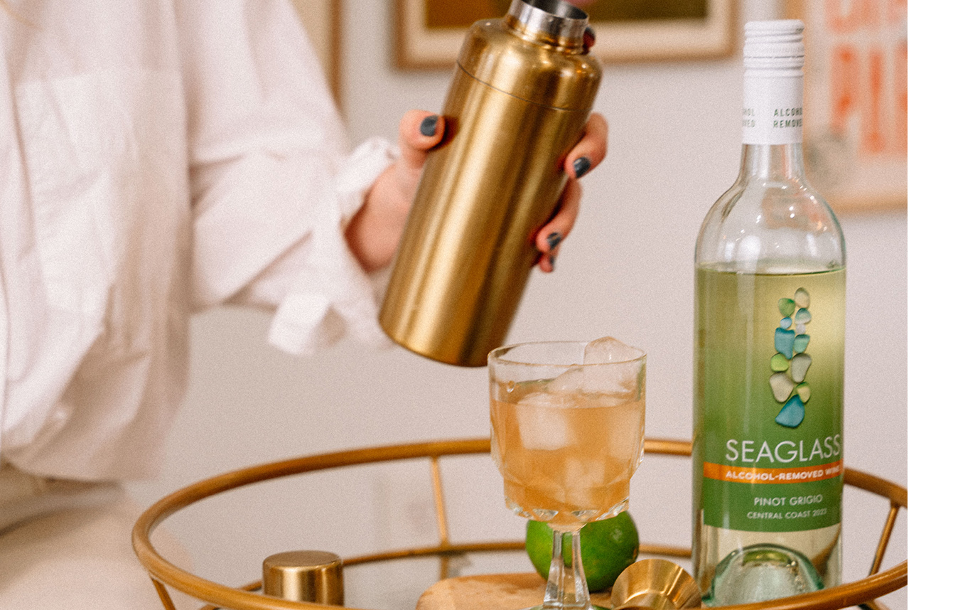 Woman in a white blouse mixing up a paloma mocktail in a gold shaker, made with SEAGLASS alcohol-removed pinot grigio. Get this mocktail recipe and more at One Stop Wine Shop.