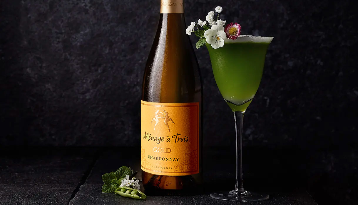 The Oh Snap cocktail, a green cocktail in a tulip glass garnished with flowers next a bottle of Menage a Trois gold chardonnay