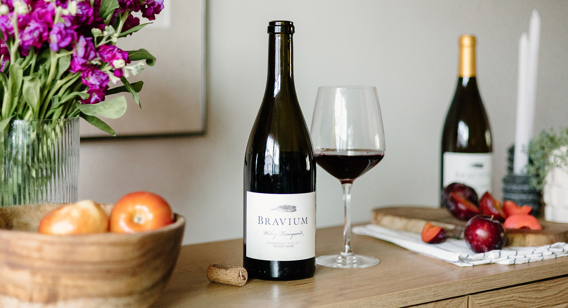 A bottle of Bravium Wiley Vineyards Pinot Noir on a wooden table next to a bowl of apples, a vase of purple flowers, and a cutting board with a sliced up plum. Get this season favorite and more at One Stop Wine Shop.