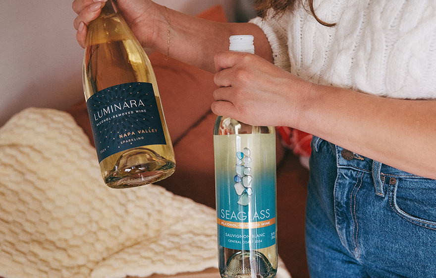 A woman in a white sweater and blue jeans holding a bottle of SEAGLASS alcohol-removed sauvignon blanc and a bottle of Luminara alcohol removed Napa Valley Sparkling.
