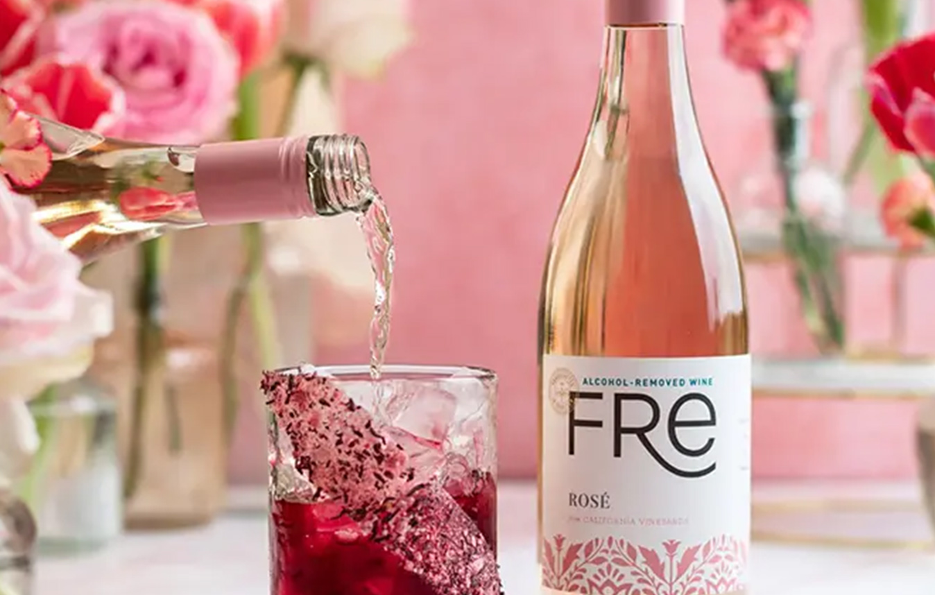 A glass of FRE alcohol-removed rose cranberry cooler, decorated with pink dried hibiscus, with pink flowers in the background. Get this mocktail recipe and more from One Stop Wine Shop