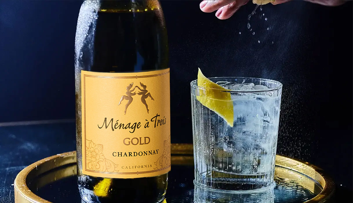 a person squeezing a lemon into a cocktail glass next to a bottle of Menage a Trois gold chardonnay