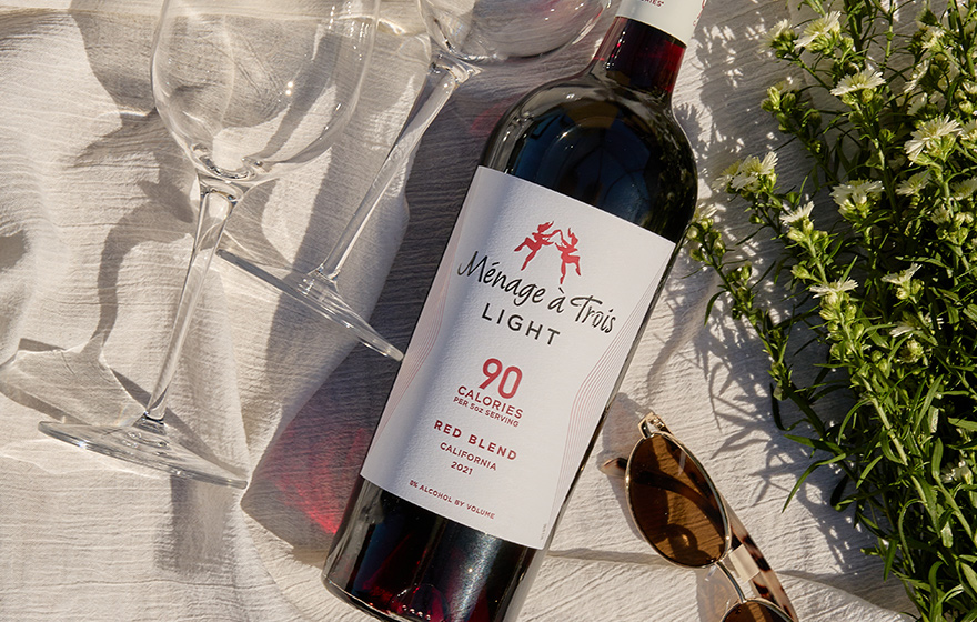 a bottle of Menage a trois light red blend, 90 calories per serving, laying on a white blanket next to two wine glasses, a pair of sunglasses, and some stalks of small white flowers.
