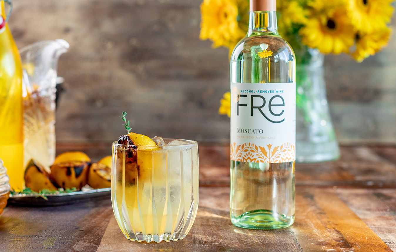A glass of smoked honey lemonade mocktail next to a glass of FRE alcohol-removed moscato with a vase of yellow flowers in the background and charred lemons on a plate.