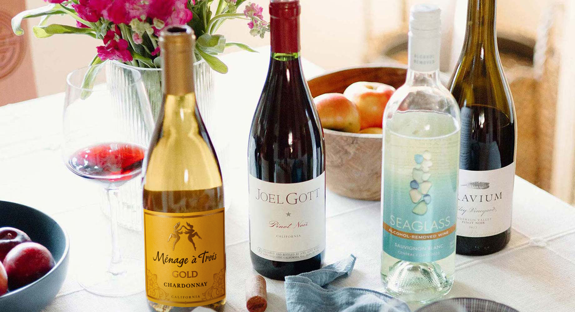 a collection of four wines on a table set with pink spring flowers and bowls of fruit. Shop our variety of exceptional wines only at One Stop Wine Shop.