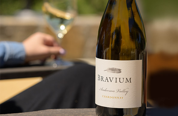 A bottle of Bravium Anderson Valley chardonnay on an outdoor table with a person in the background enjoying a glass of wine. Shop this seasonal favorite and more from One Stop Wine Shop
