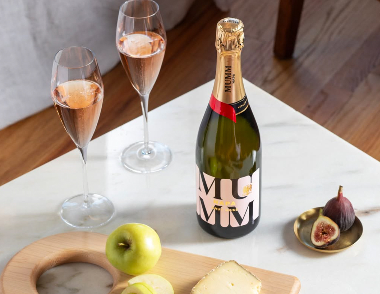 A bottle and two glasses of Mumm Napa Brut Rose on a marble countertop next to a fig and apple.