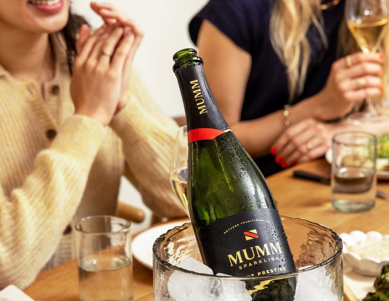 A bottle of Mumm Napa sparkling Brut Prestige chilling in a container with ice with two women in the background smiling and enjoying glasses of wine with a meal.