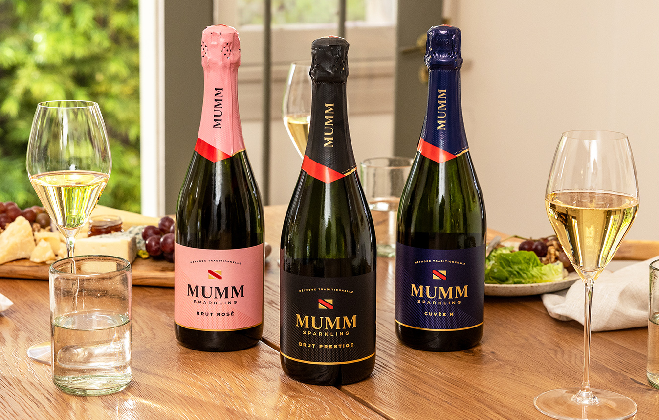 Three bottles of Mumm Napa sparkling wines on a wooden dinner table with glasses of sparkling wine around them and a cheese plate in the background. Shop now at One Stop Wine Shop.