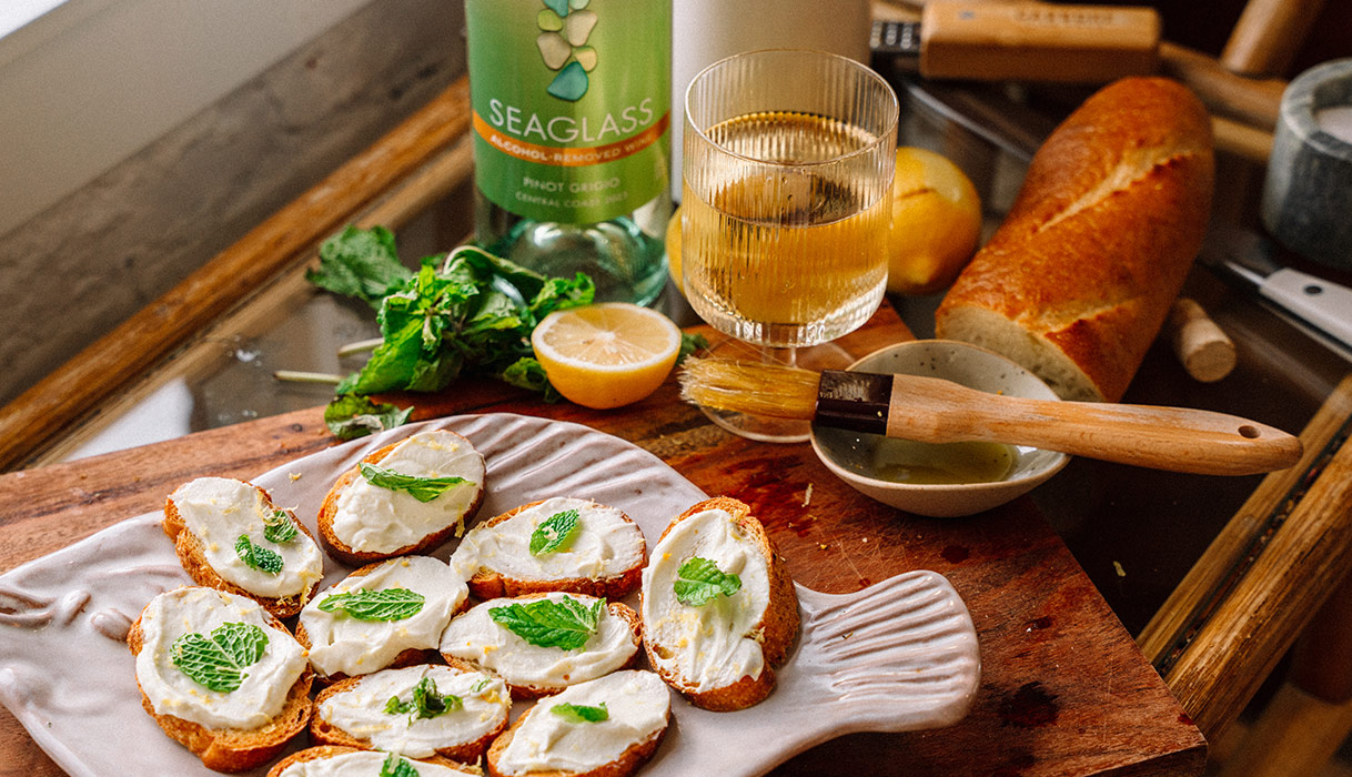 a plate of citrus whipped goat cheese crostini with a glass and bottle of SEAGLASS alcohol-removed pinot grigio in the background. Get this recipe and more at One Stop Wine Shop.