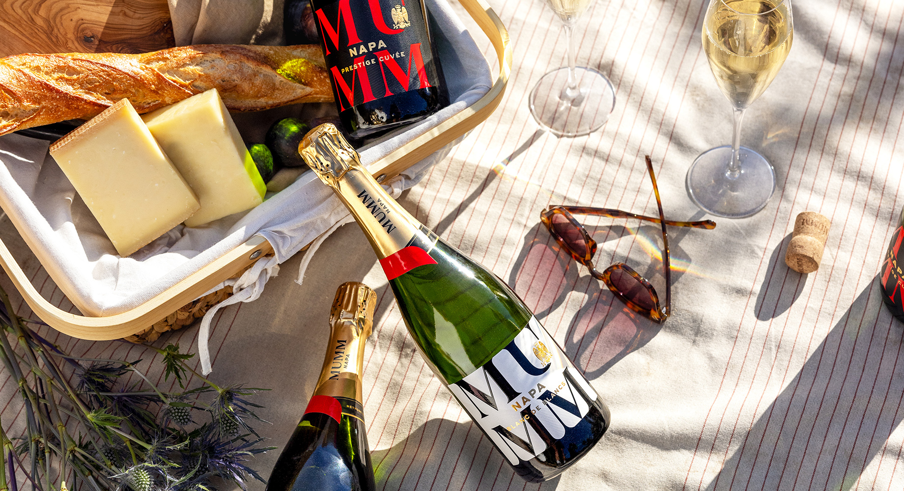 New Mumm Napa wines laying on a picnic blanket surrounded by bread, cheese, and dappled with sunlight. Shop these and more new arrivals at One Stop Wine Shop.