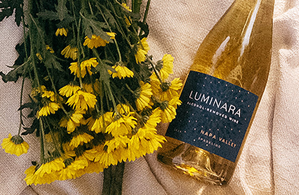 a bottle of Luminara Alcohol-Removed Napa Valley Sparkling next to a bouquet of yellow flowers on a blanket. Shop our wide selection of alcohol-removed wines at One Stop Wine Shop.