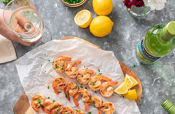 lemon butter shrimp skewers on a wood cutting board surrounded by sliced lemons and bottles of Pomelo sauvignon blanc. Get this recipe and more at One Stop Wine Shop.