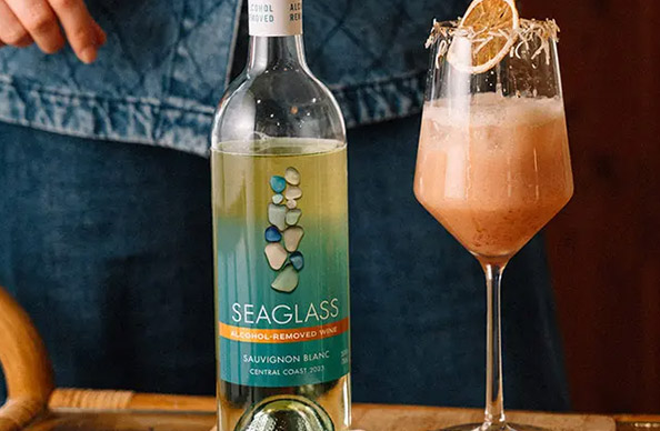 a bottle of SEAGLASS alcohol-removed sauvignon blanc next to a coconut blood orange mocktail garnished with toasted coconut. Get this mocktail recipe and more at One Stop Wine Shop.