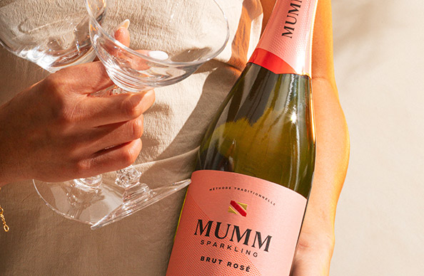 Woman in a linen shirt holding wine glasses and a bottle of Mumm Sparkling Brut Rose. Shop this sparkling wine and more at One Stop Wine Shop.