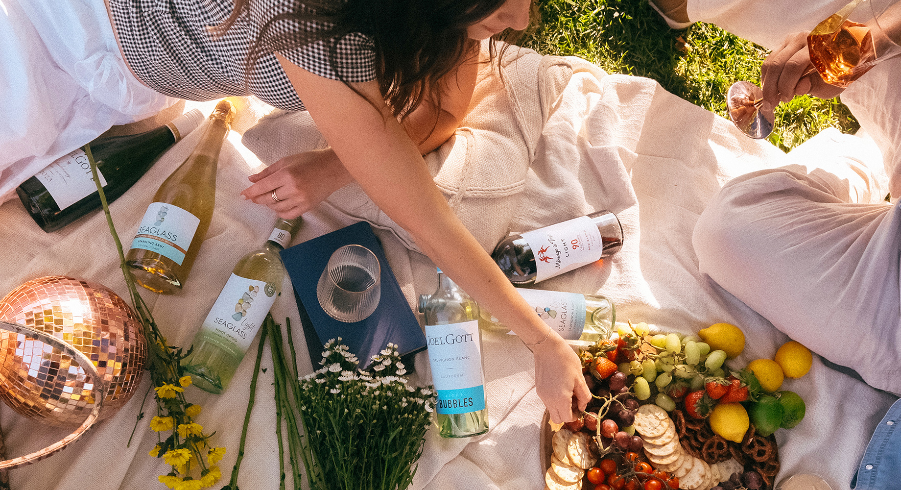 People enjoying a variety of seasonal One Stop Wine Shop wines on a cream colored picnic blanket surrounded by snacks and bouquets of flowers. Shop our curated selection of seasonal wines at One Stop Wine Shop.