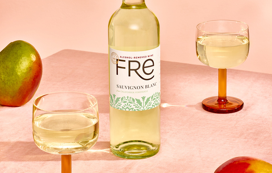 A bottle and glasses of FRE alcohol-removed sauvignon blanc with a light pink background and mangos. Shop our wide selection of alcohol-removed wines at One Stop Wine Shop.
