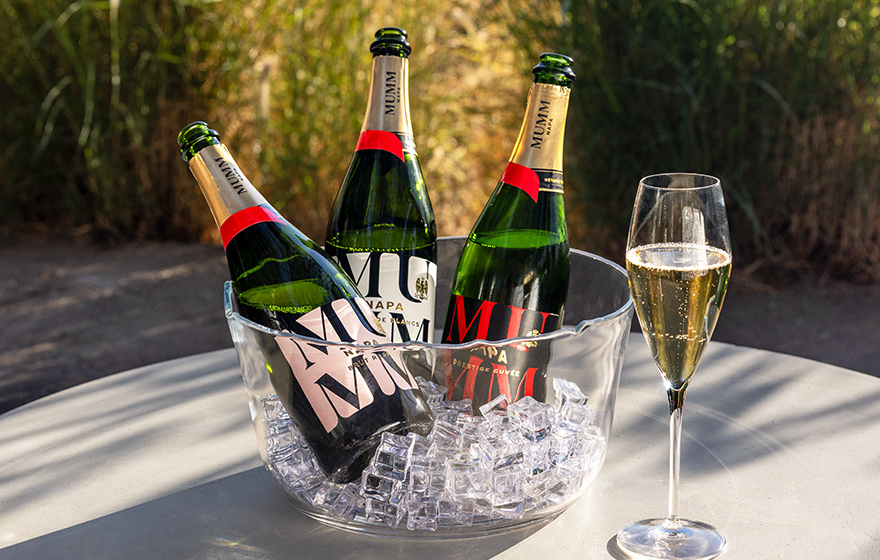 Bottles of Mumm Napa wines chilling over ice in a glass container with lush greenery in the background. Shop Mumm Napa at One Stop Wine Shop.