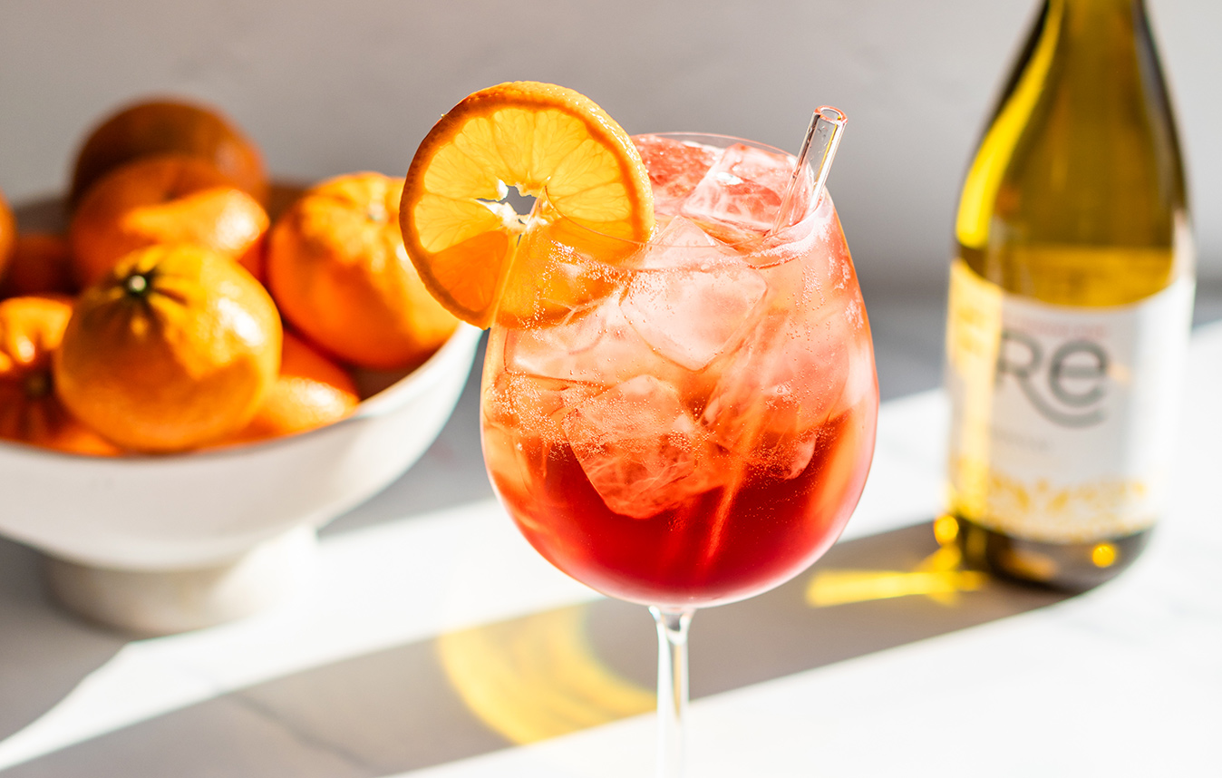 A vibrant orange glass of classic Italian spritz mocktail made with FRE alcohol-removed Chardonnay with a bowl of oranges in the background. Get this mocktail recipe and more at One Stop Wine Shop.
