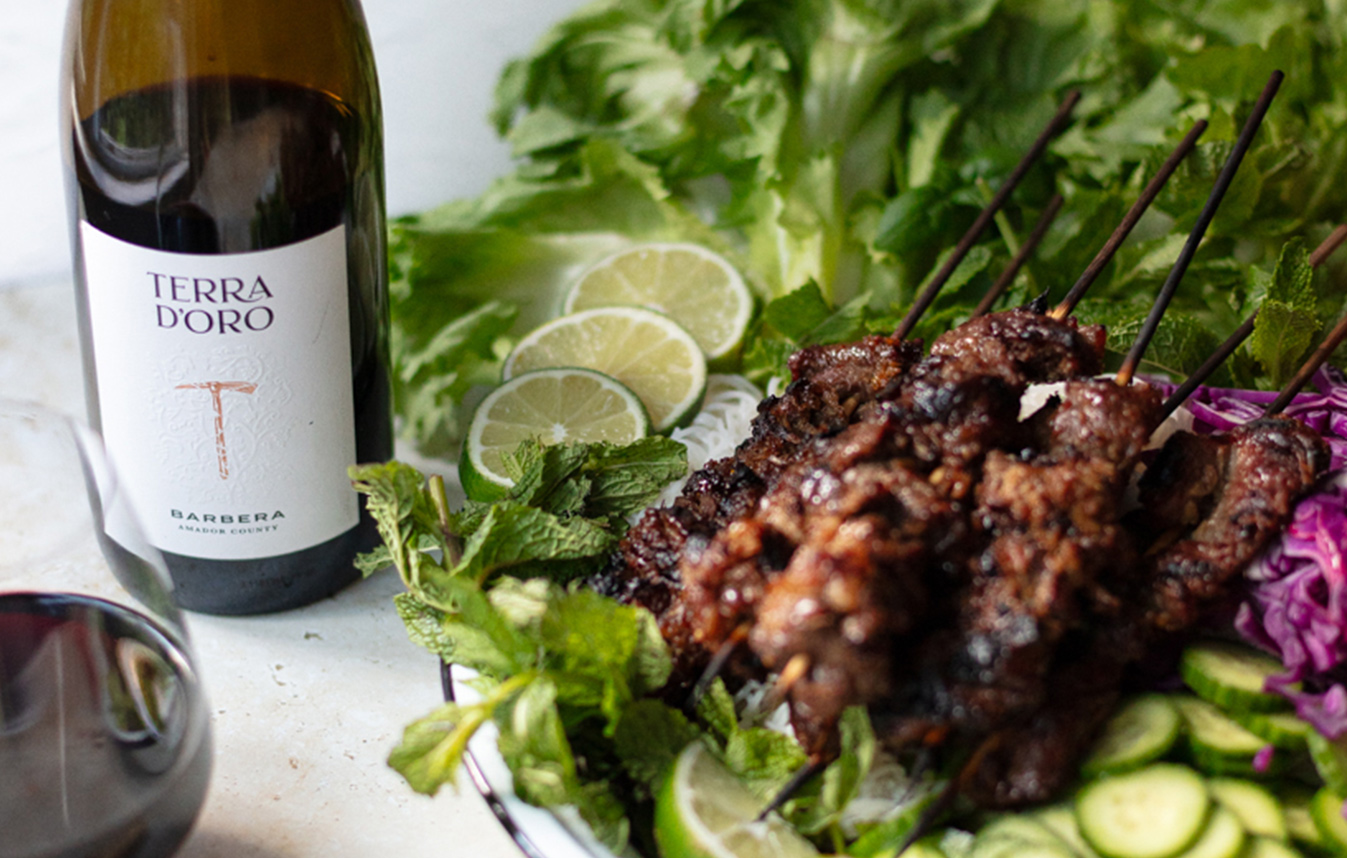 a plate of lemongrass beef skewers surrounded by sliced limes, cucumber, and lettuce, next to a bottle and glass of Terra d'Oro Barbera.