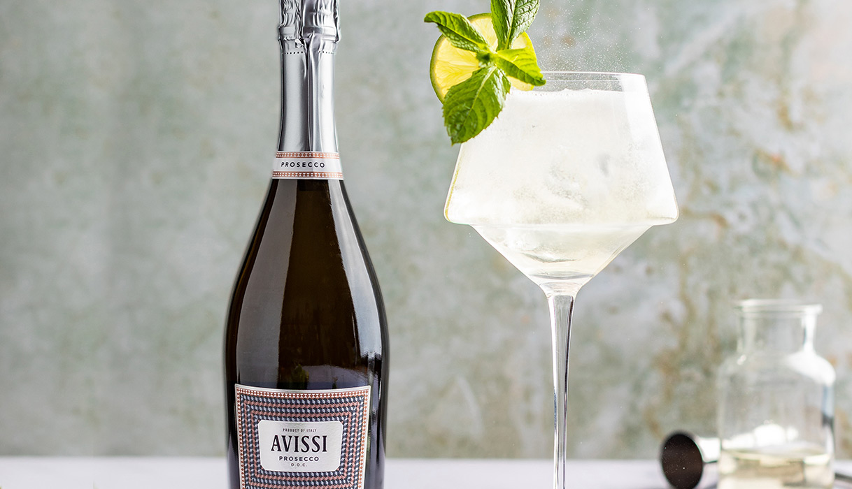 A Hugo Spritz in a fancy glass garnished with a lime wheel and mint with bottles of Avissi Prosecco in the background. Get this cocktail recipe and more from One Stop Wine Shop.