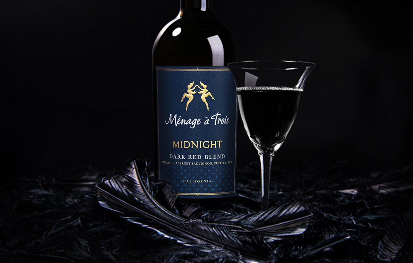 A bottle of Menage a Trois midnight dark red blend next to a glass of wine with dark black feathers underneath