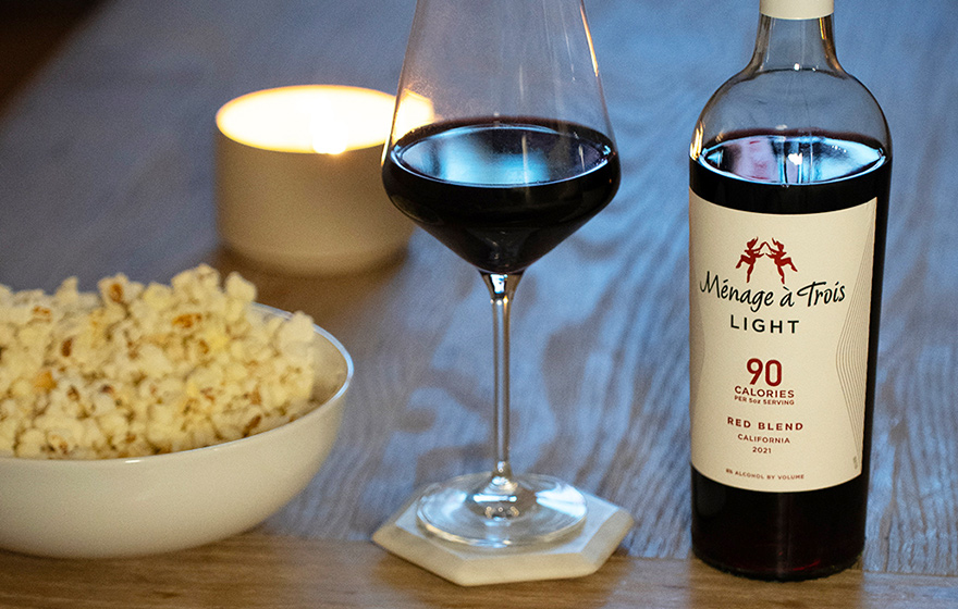 A bottle of Menage a Trois light red blend next to a glass of wine, a bowl of popcorn and a light candle