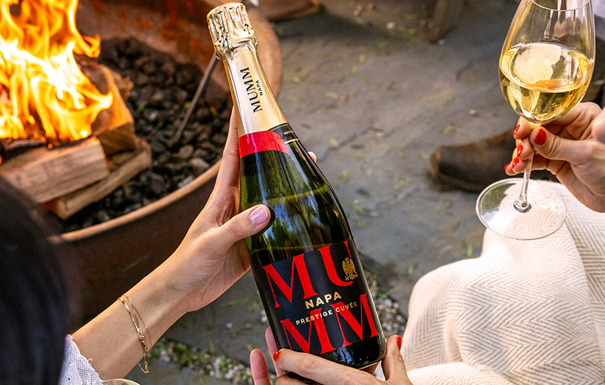 Two woman looking at a bottle of Mumm Napa prestige cuvee next to a fire pit