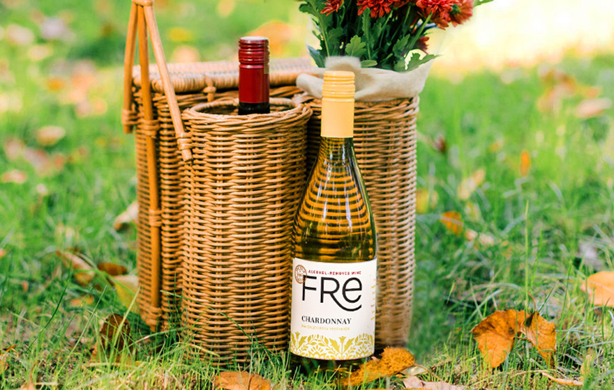 A bottle of FRE alcohol-removed chardonnay next to a basket with another bottle of FRE wine and mumms all in the grass with fall leaves around it