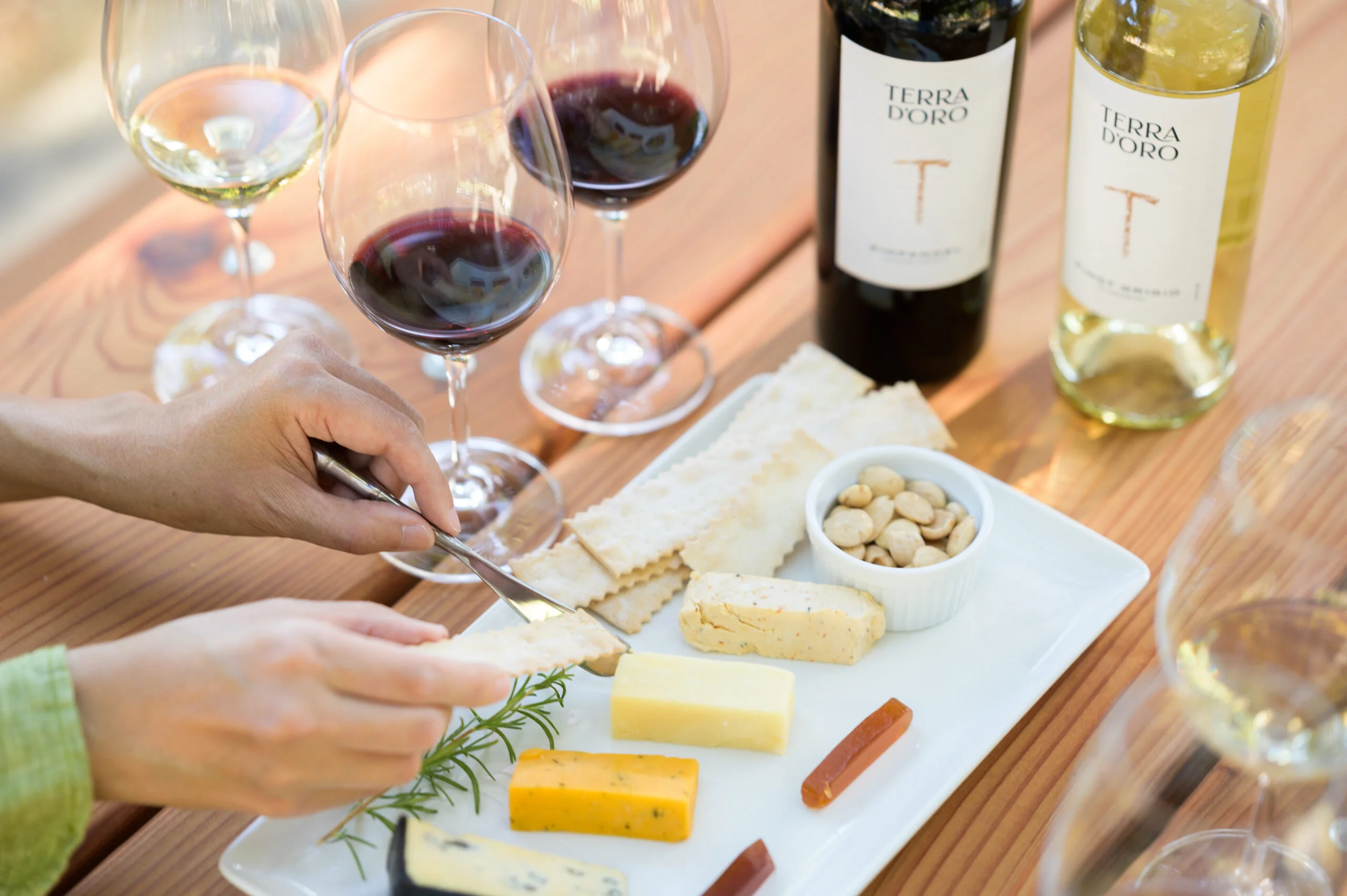A woman places some cheese on a cracker next to a platter featuring various cheeses, jams, and nuts alongside bottles of Terra d'Oro red and white wines
